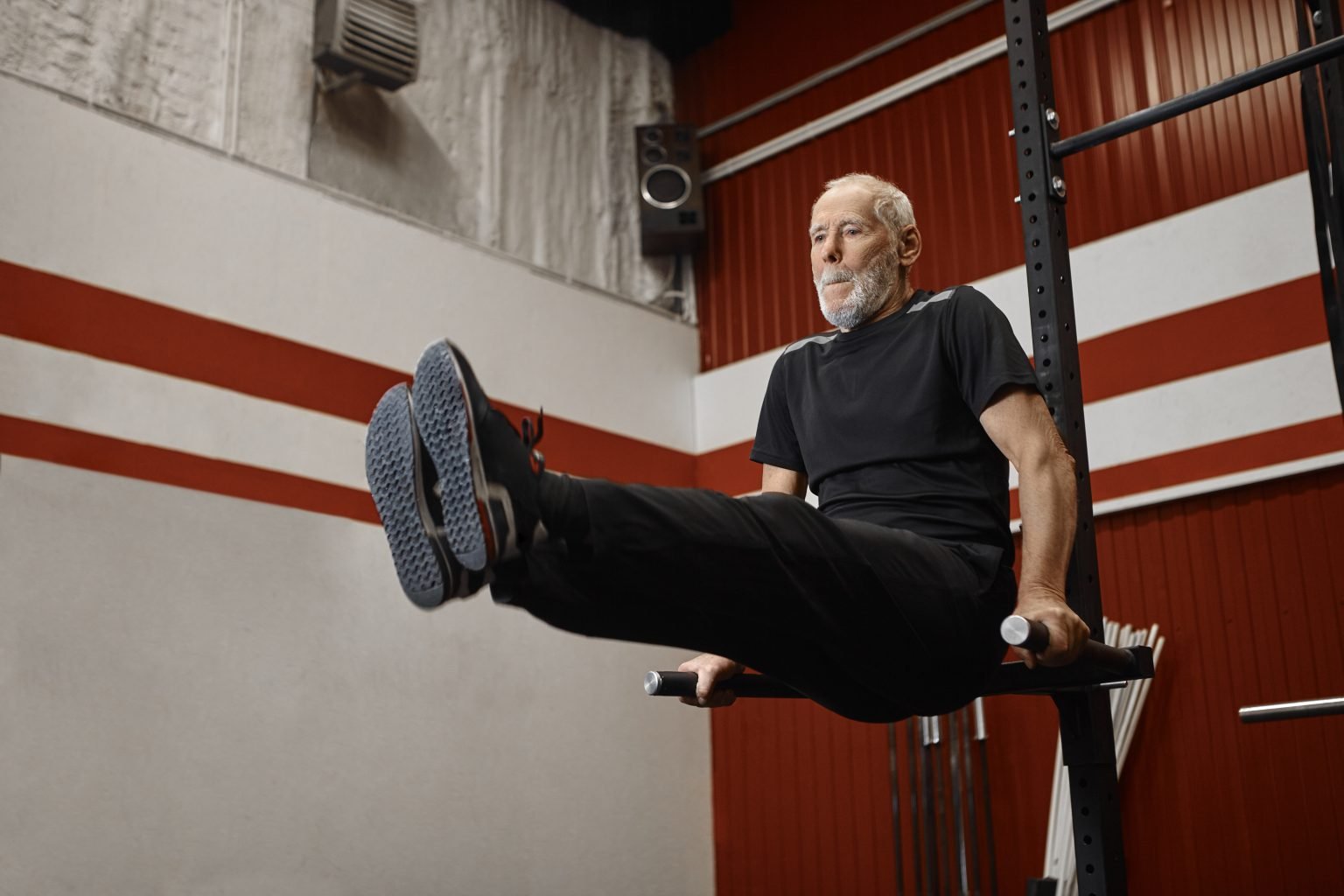 Handsome usnhaven retired man in stylish sportswear doing crossfit training in gym, raising legs while exercising on pull up bar, strengthening abdominals. Fitness, sports and retirement concept